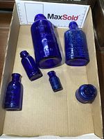 Seven cobalt blue glass bottles arranged in a cardboard box with measurement scale showing their heights and varying sizes.