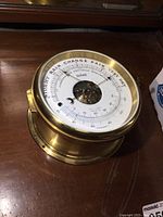Front and angled front view of vintage brass ships barometer on wooden surface showing dial and thermometer scale.