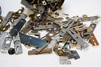 Overview of assorted metal door hinges, strike plates, knobs, and other small hardware items spilled from a tin container on a white surface.
