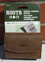 Front view of the brown Roots digital camera pouch in packaging showing model RAF10H and product name.