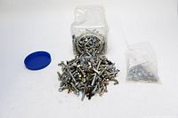 Two piles of mixed screws poured from a plastic container and a smaller plastic bag on a white surface