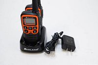 Front view of one Midland X-Talker walkie talkie in black and orange placed in Midland branded charging dock with power adapter cable coiled next to dock