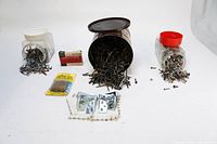 Three jars containing a variety of mixed screws, nails, metal fasteners, corner brackets, and chains spread on white background