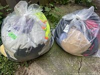 Two large clear plastic bags filled with assorted mixed clothing including visible neon yellow garment, beige, black and other colored clothing items, placed on outdoor ground.