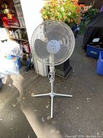Full view of the white floor fan standing outdoors with a tripodal base and a long power cord wrapped around the pole.