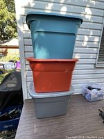 Stack of three Sterilite storage bins in blue, orange, and gray shown outdoors beside house wall.