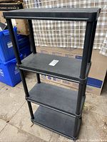 Black 4-shelf plastic shelving unit, full view showing all shelves and legs, placed indoors near cardboard boxes and plastic containers.