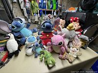 Photo shows a variety of Disney stuffed toys including multiple Stitch plushes in various sizes, Minnie Mouse, Dumbo, and other assorted plush animals arranged on a table.