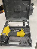 Yellow and black air stapler/nailer tool inside a black plastic carrying case, along with staples and some accessories, showing dust and wear.