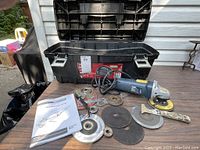 Angle grinder with case, multiple grinding and cutting discs, wrench, and instruction manual displayed on wooden surface outside.