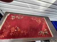 Rectangular handmade Chinese rug with a red background and floral designs placed on a wooden surface.