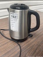 Stainless steel electric kettle with black plastic handle, lid, and base, plugged in, shown from an angle.