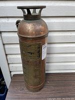 Full view of the antique fire extinguisher showing copper/brass body and top handle structure on wooden surface.