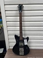 Full view of the black First Act electric guitar leaning against white siding. Shows the body, neck, and headstock with tuning pegs.