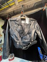 Front view of the black leather jacket showing the zippered pockets, shoulder epaulets, and belted waist with metal buckle.