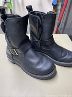 Side and front view of both boots showing black leather material, buckle strap, and overall wear condition.