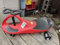 Side view of PlasmaCar kids' ride-on toy car showing red body, black steering wheel, seat and footrests.