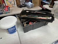 Black plastic toolbox open with visible assorted hand tools inside, next to the white bucket of drywall compound on a white table.