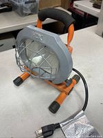 Front view of single orange halogen work light with gray protective metal cage and cushioned black handle showing the lamp glass.
