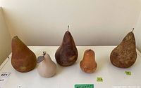 Front view of all five decorative pears lined up side by side on a flat white surface, shows size differences and material textures