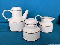 Front angled view of tea pot, milk jug, and sugar bowl on blue background showing stoneware texture and dark rim detailing