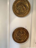 Photo of two brass plates hanging vertically on a white wall, showing their tavern scene designs and ornate borders.