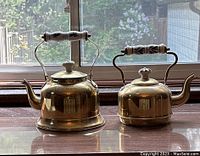 Photo showing both brass teapots side by side on window ledge with natural light. Displays size difference and handles.