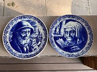 Two blue and white Delft ceramic plates placed side-by-side, showing the elderly man and woman designs with floral borders on the edges.