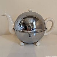 Front view of vintage Ever-Hot teapot showing shiny chrome outer dome, ceramic handle, spout, and lid knob, resting on three ceramic feet.
