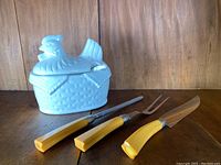 Three-piece vintage Bakelite handle carving set comprising knife, fork, and sharpening steel, displayed next to a ceramic hen and basket-shaped soup tureen with lid.