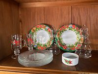Overview of two plastic serving bowls with holly and berries decoration, four clear glass Christmas tree plates, two clear glass tree decorations, two glass footed mugs with Christmas holly design, and a small ceramic ramekin.