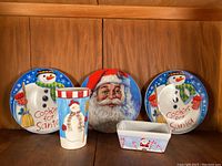 Photo of four Christmas themed dishes: two plates with snowmen and 'Cookies for Santa' text, one cup with snowman illustration, one rectangular dish with Santa and candy canes.