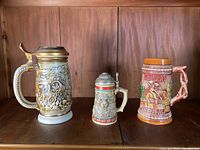 Front view of three ceramic decorative steins on shelf, showing Avon Gold Rush, Mini Jaguar, and California Redwoods themes with relief designs and lids.