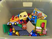 View inside a half tote showing a variety of Duplo blocks including base plates, a yellow carriage, vehicles, and animal figures.