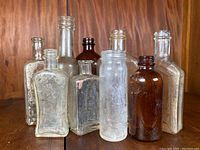 Nine vintage bottles and jars in varying sizes, colors, and shapes arranged in front of wood panel background