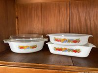 Four pieces of white glass bakeware with colorful fruit pattern arranged on wooden shelf, showing the set and clear glass lid.