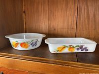 Two Fire King bakeware dishes, round and rectangular, set on wood surface with fruit design visible.