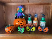 Display of Halloween decor items on wooden shelf including ceramic jack-o-lanterns, three potion jars, three pairs of Halloween eye glasses, and a stuffed witch doll.