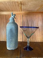 Front view of vintage seltzer bottle with metal woven mesh sleeve next to large glass martini glass with blue rim and base on wooden shelf.
