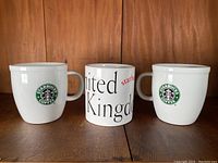 Front view of three vintage Starbucks mugs on wooden surface, showing early logo on two mugs and UK telephone booth design on the third