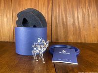 Photo showing the Swarovski crystal fawn figurine standing next to its blue cylindrical box with custom black foam insert and the certificate booklet.