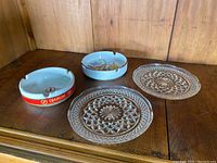 Four items on wooden surface: two white ceramic ashtrays with red Djarum band, two small clear decorative glass plates with cut patterns.