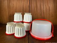 Photo of six white Tupperware dessert molds with red bases and lids, plus a mini cake saver with red handle and base, set against a wood background.