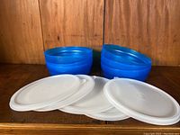 Six blue stacking bowls arranged in two stacks, with six white lids placed in front of them on a wooden surface.