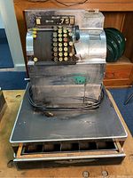 Front view of vintage National cash register showing keypad, mechanical display, crank mechanism side, and metal body on black base.
