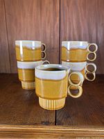Four vintage ceramic coffee cups with two-finger ring handles arranged in pairs on a wooden surface with a warm mustard and cream glaze.