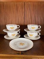 Four white ceramic cups and six matching saucers, all with wheat pattern and gold rim, arranged on wooden surface.
