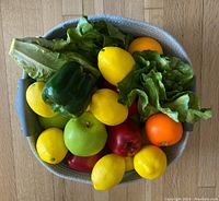 Basket filled with artificial fruits and vegetables including green bell pepper, yellow lemons, green apple, red apples, orange, lettuce, and a red pomegranate, showing overall contents in the basket.