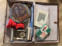 Open vintage Bukowski briefcase showing interior items including books, decorative plate, red shoe figurines, clock, and lighter