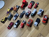 Top and side view of 18 diecast toy vehicles arranged on wooden floor showing various trucks, tractors, racing cars and tankers.
